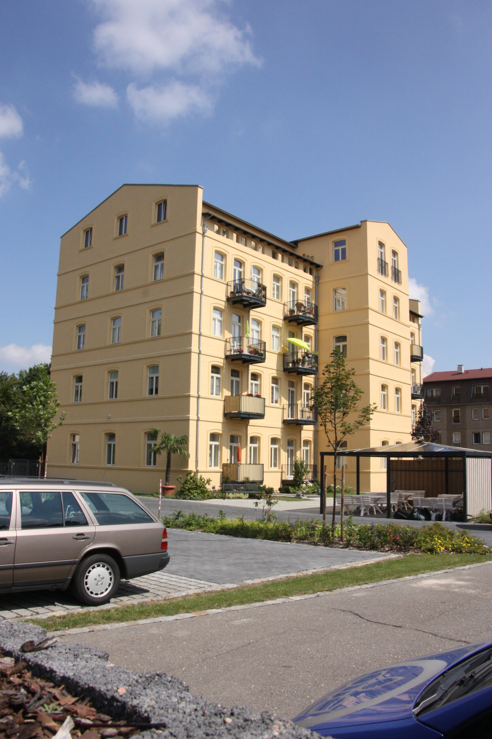 Gelbes Mehrfamilienhaus mit Balkonen, Parkplatz und Grünfläche. Planungsbüro.